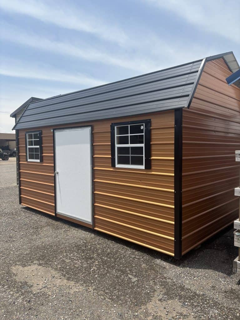Metal storage shed with brown siding and black trim, featuring two windows and a white door, set on gravel ground under a blue sky, showcasing GEMCO Buildings, Inc. metal building construction services.