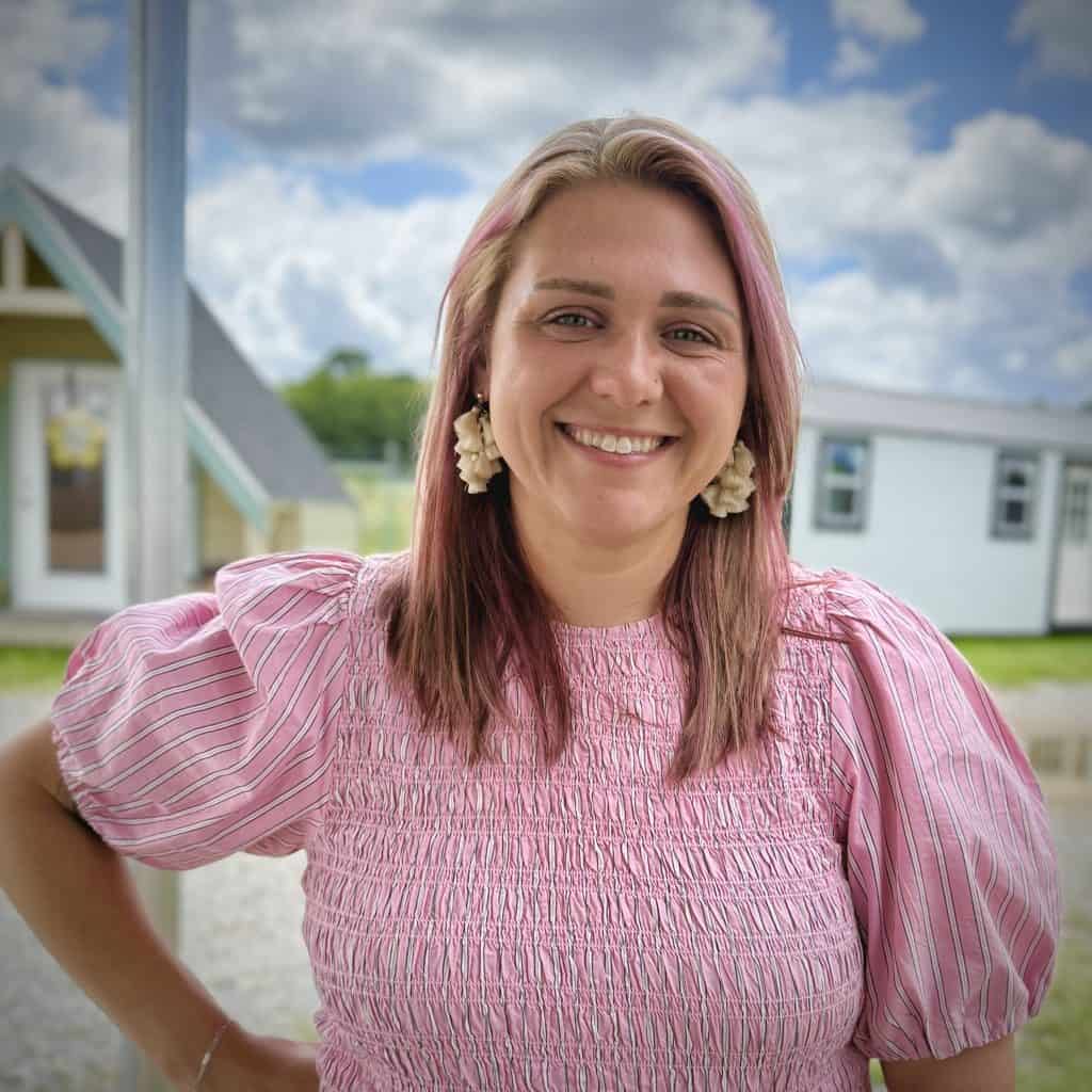 Smiling woman with pink hair and floral earrings standing outdoors in front of GEMCO Buildings, Inc. location during daytime.